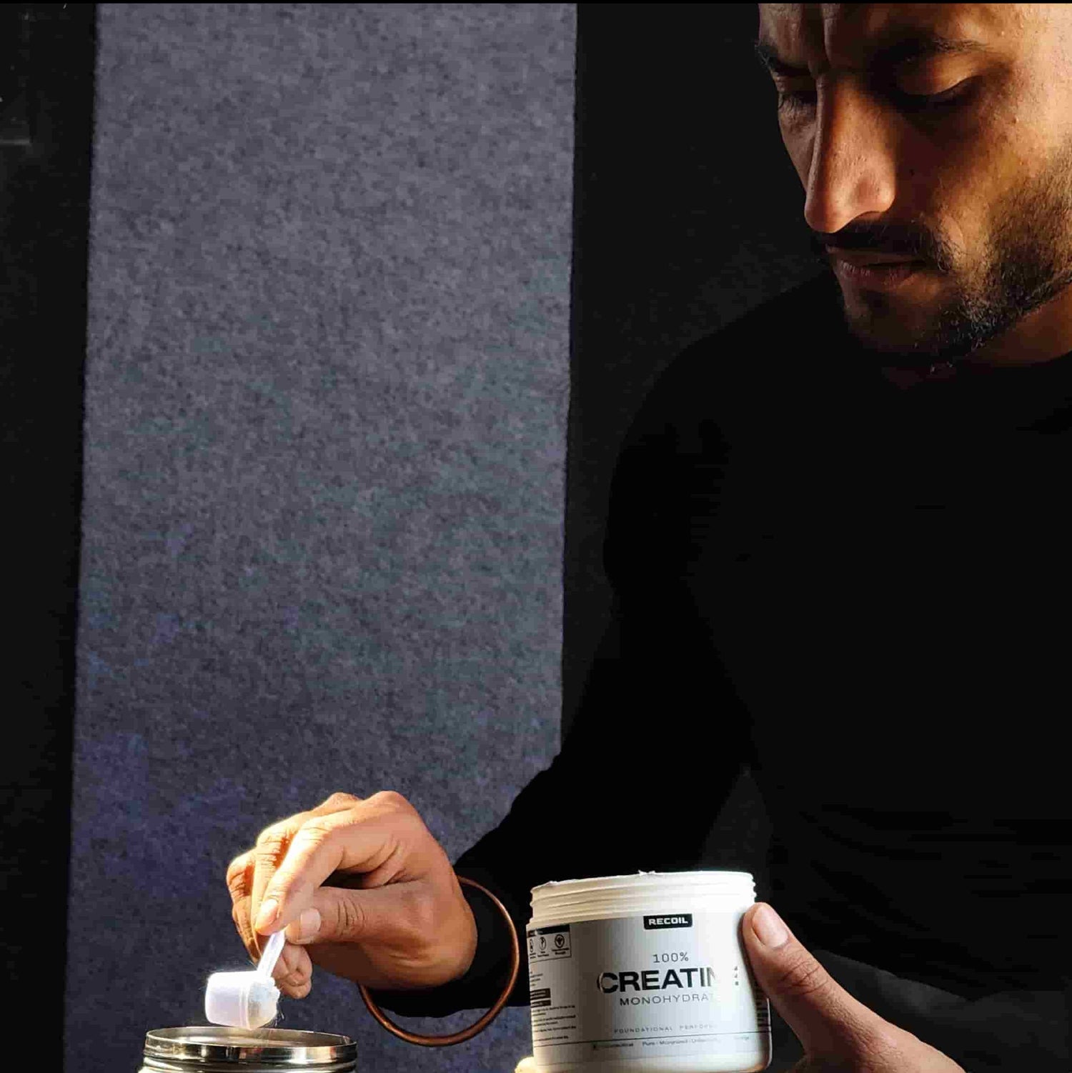 Person holding a container labeled recoil  'CREATine' next to a metal cup against a dark background